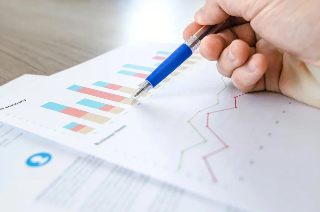 A close-up of a person's hand using a blue pen to point at printed financial charts and line graphs.